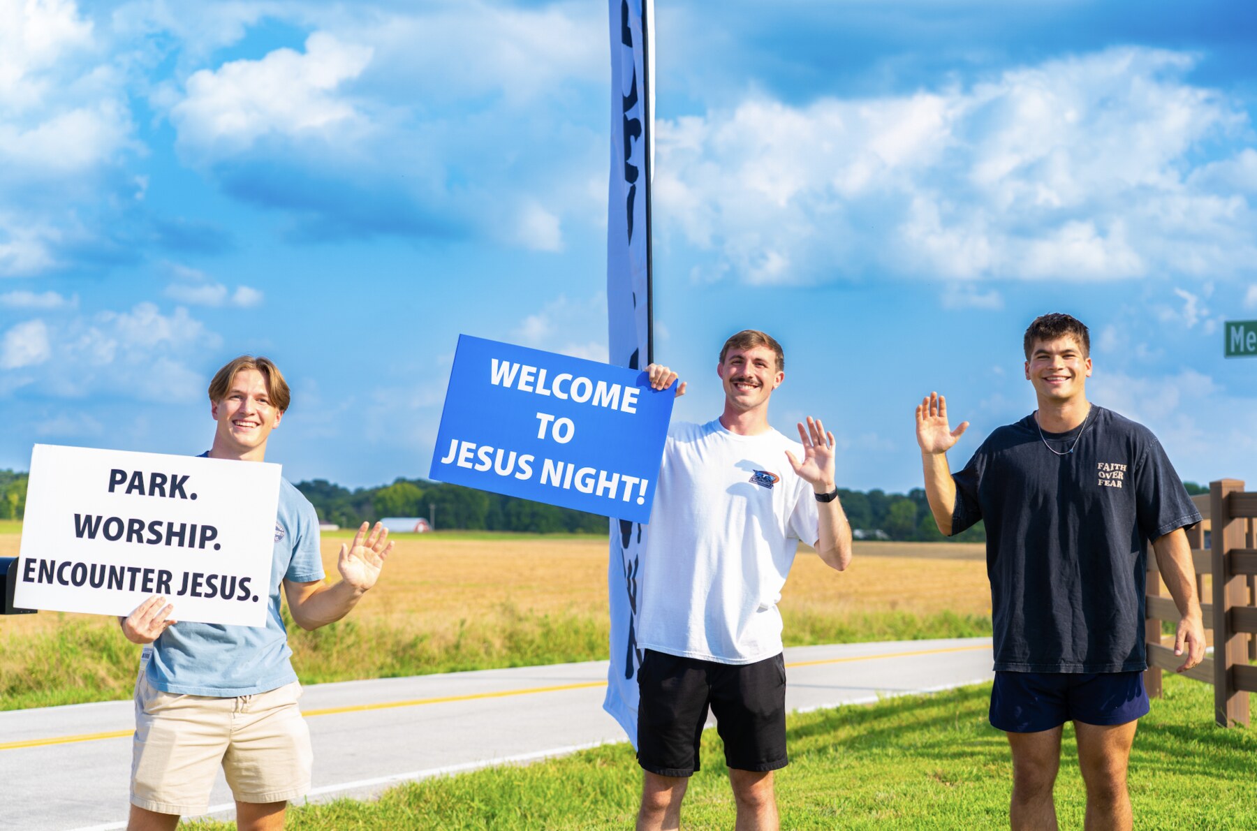 Three volunteers waving and holding signs welcoming people to Jesus Night