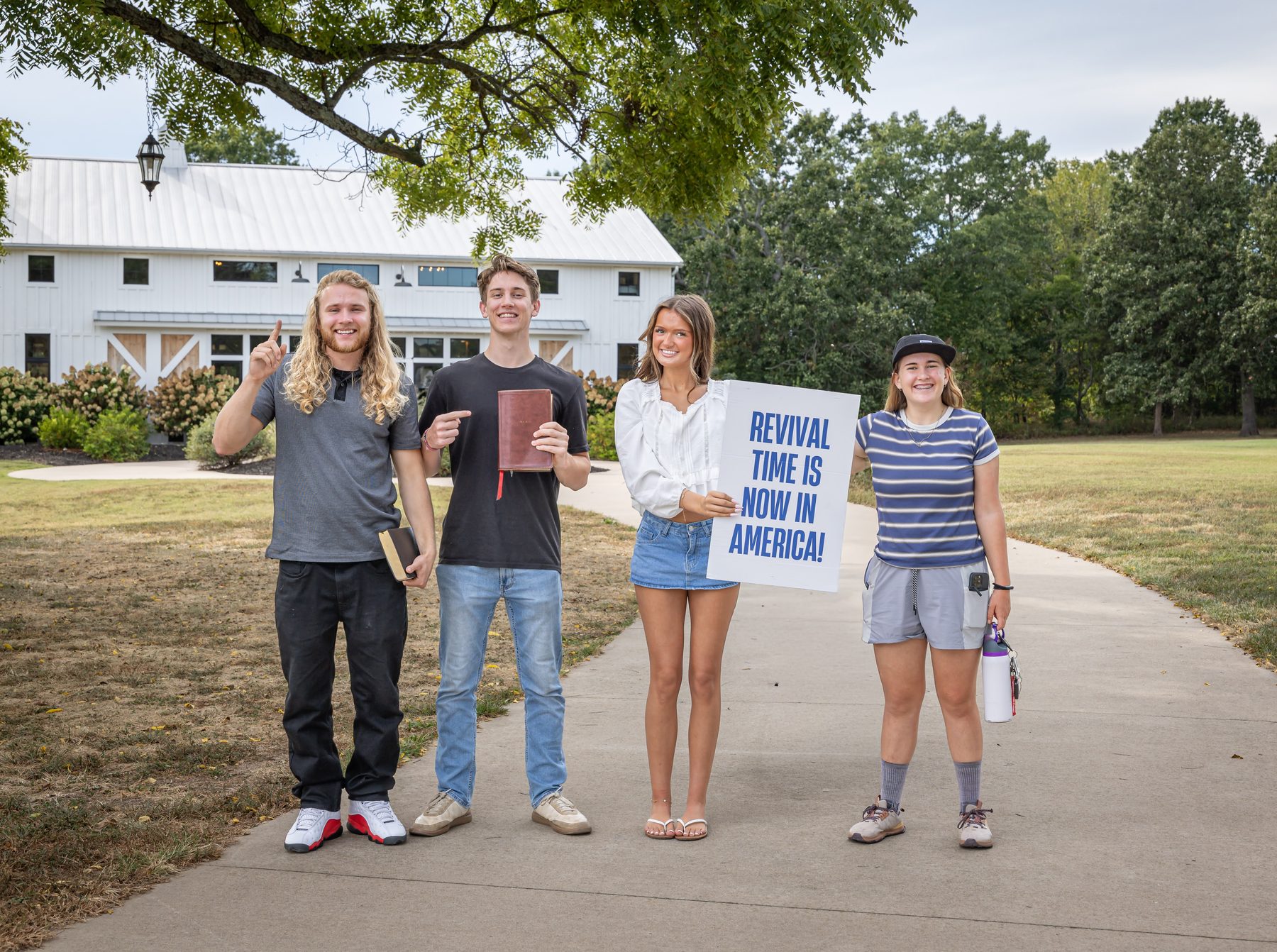 Four young adults standing on the walkway outside the Jesus Place Missouri barn, holding Bibles and a sign that reads "Revival time is now in America!"
