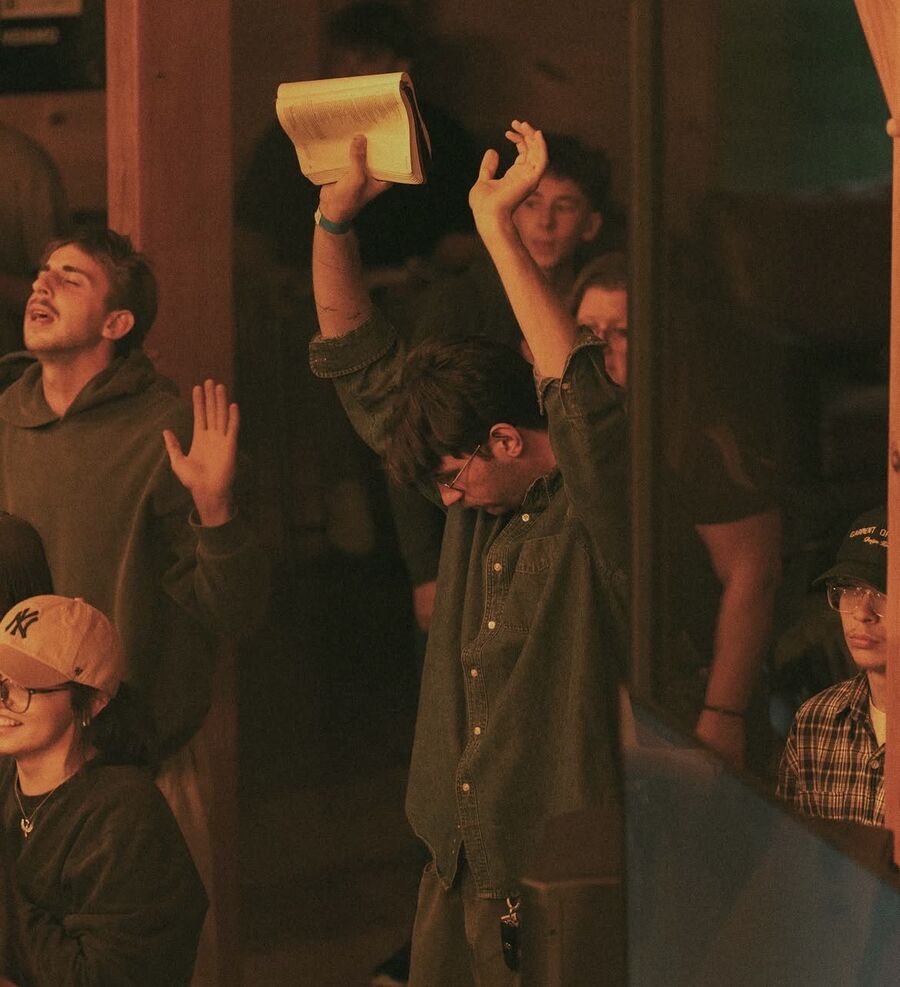 A young man lifts an open Bible high with both arms raised in worship while others around him sing