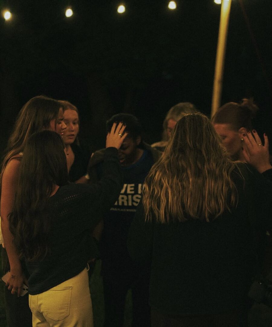 A group of friends gathers under string lights at night to lay hands on and pray over someone
