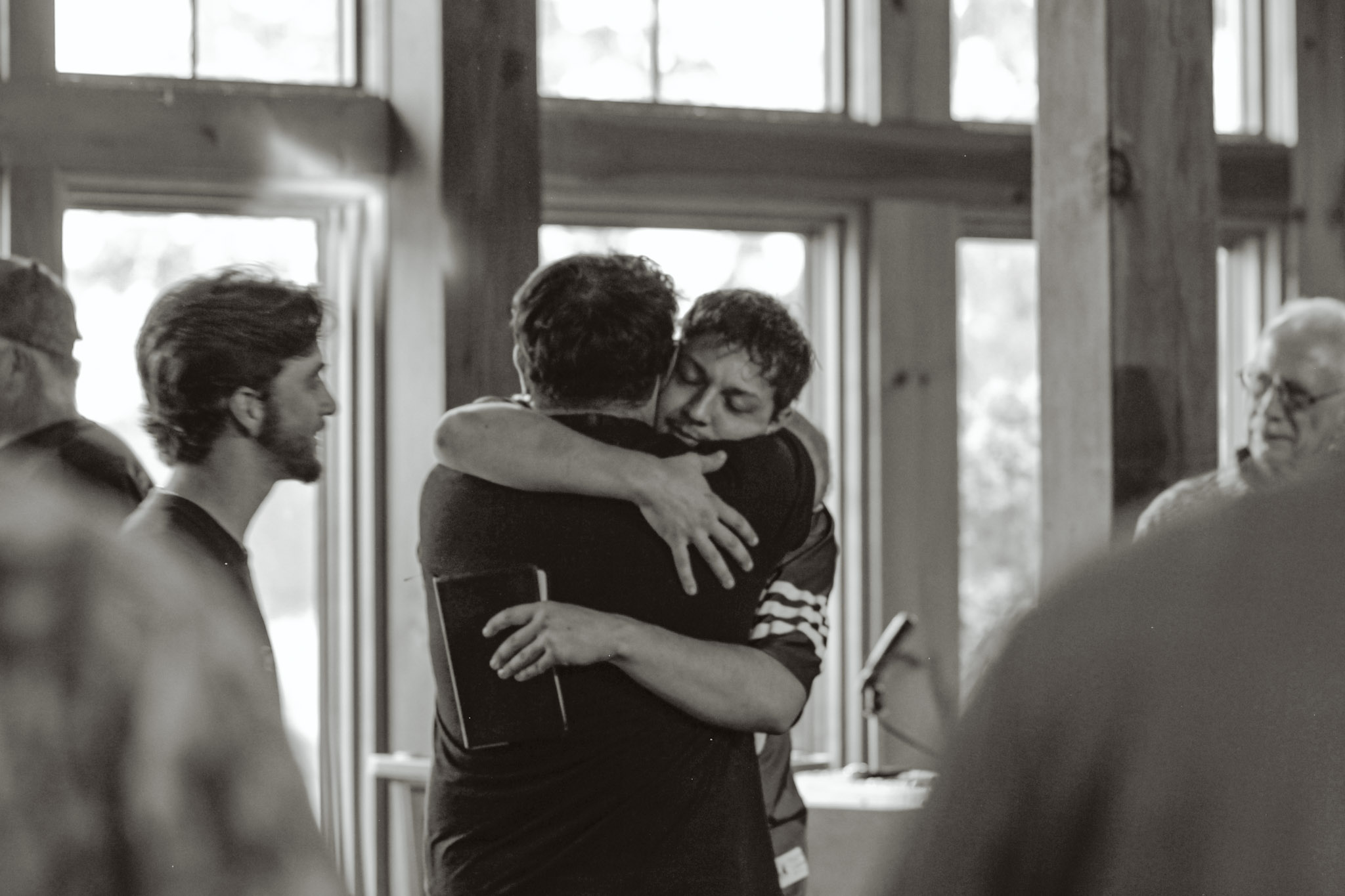 Two young men sharing a long embrace, one holding a Bible, as friends look on in the lodge