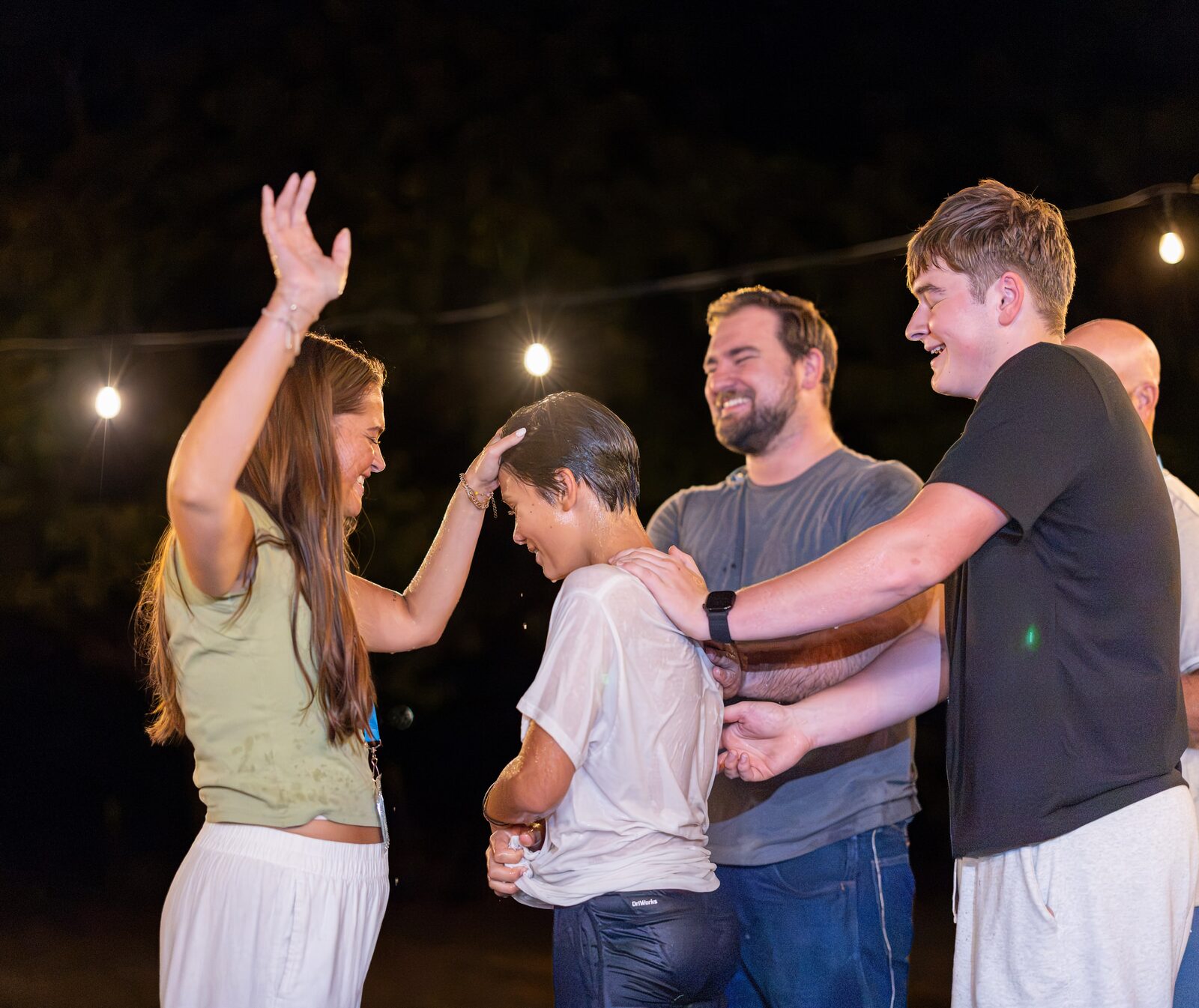 Friends laying hands on and praying over someone just baptized, outdoors under string lights