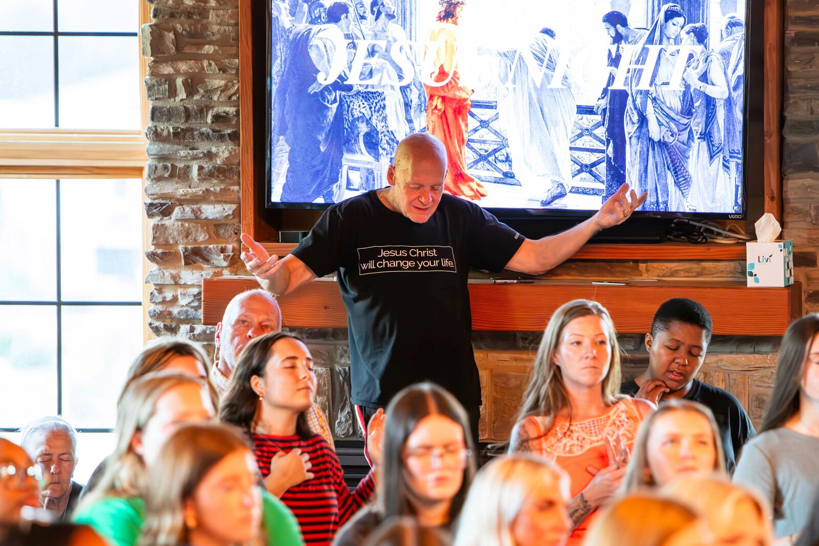 A speaker with arms outstretched praying over the gathered room in front of a stone fireplace