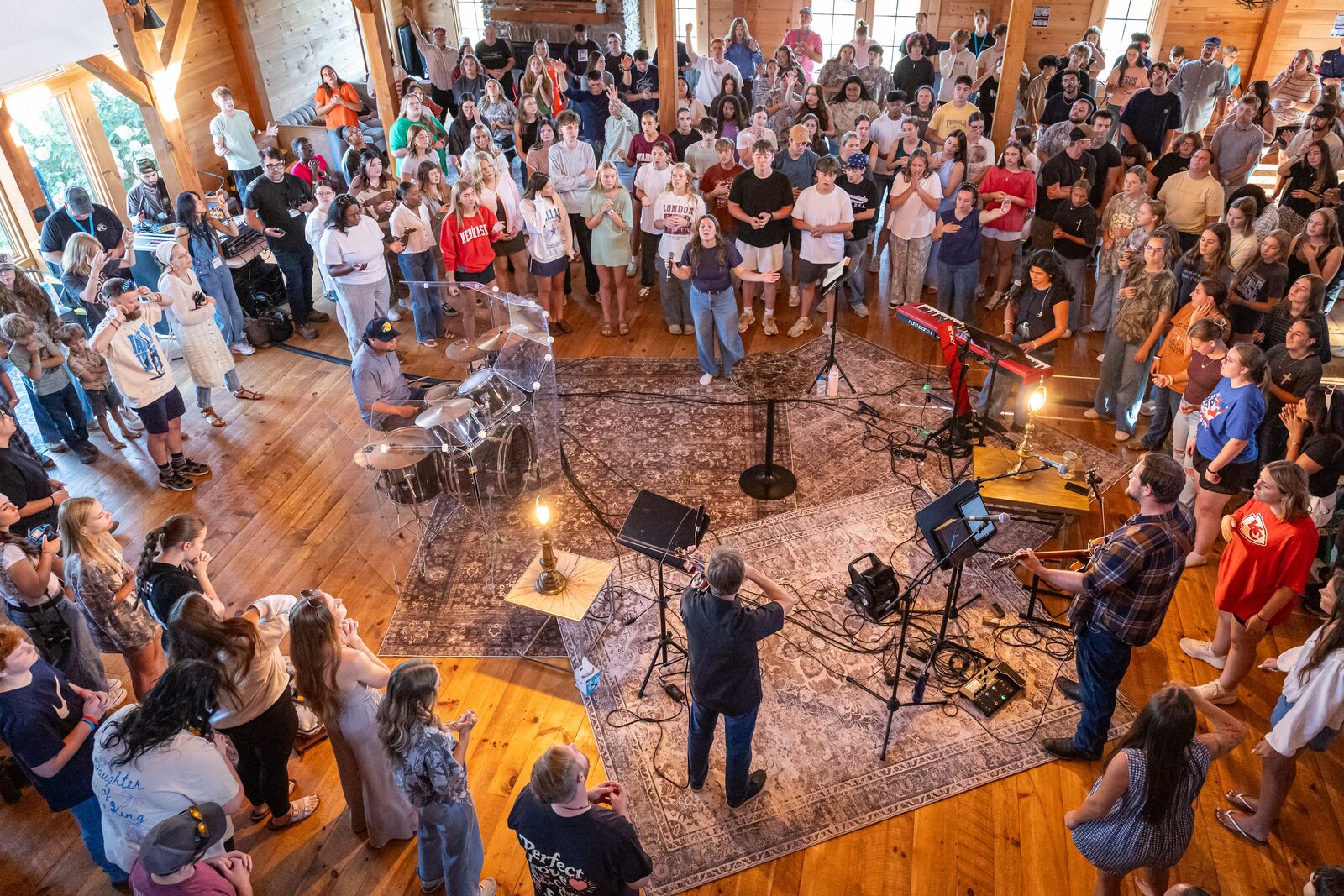 Black-and-white overhead view of a packed worship gathering surrounding a live band on rugs in a wood-paneled hall